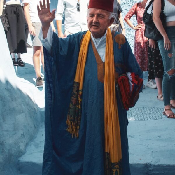 An elder in traditional Moroccan clothing waves in a bustling Chefchaouen street.