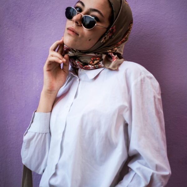 Chic woman in hijab and sunglasses posing against a vibrant purple wall, showcasing modern fashion.